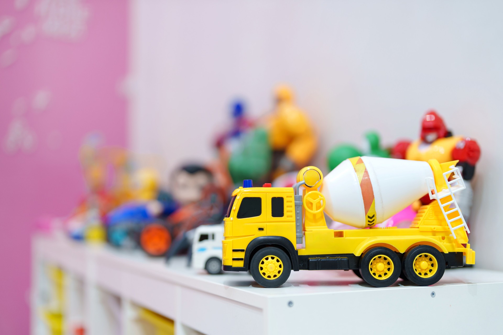 Various colorful toys are neatly arranged on a storage cabinet in a kid's playroom, with the image focusing on a yellow concrete truck mixer toy.